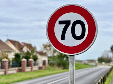 Kırsal bir yol kenarında, kırmızı kenarı ve beyaz arka planı olan, bulutlu bir gökyüzü altında bulanık evler ve yeşilliklerin bulunduğu, trafik kurallarını, trafik kurallarını, sürüş güvenliğini ve uyarıyı simgeleyen, yuvarlak bir 70 km / saat 'lik yol işaretine yakın çekim