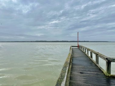 Le Crotoy ahşap iskelesi Somme Körfezi üzerinde kırmızı fener, gelgit suları ve yumuşak bulutlu gökyüzü ile sakin gri bir günde Saint-Valery-sur-Somme 'ye doğru geniş bir kıyı manzarası