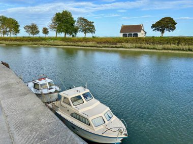 Saint-Valery-sur-Somme, Fransa - 17 Ekim 2025: Quai Blavet, kaldırımlı rıhtım boyunca demirlemiş iki tekne ile nehir kenarındaki beyaz bir kulübe ve gelgit kanalı yanında rüzgar şeklinde ağaçlar.