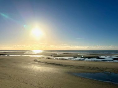 Quend ve Fort-Mahon arasındaki engin gelgit sahillerinde yürüyen iki kişi, gelgit havuzları ve yumuşak altın gün batımı gökyüzü altında güneş ışığı yansımaları.