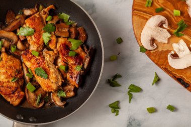 İçinde tavuk göğsü sote olan çubuksuz kızartma tavasının düz görüntüsü. Et otlar, domates, baharatlar, maydanoz, taze soğan ve mantarla tatlandırılır. Kesme tahtası var. 