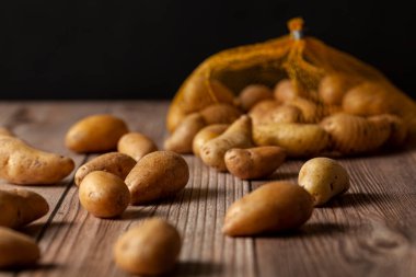 Shallow depth of field dark image of small irregular shaped ratte potatoes scattered on the surface of a wooden table from a tipped over mesh sack. An artistic versatile low light still life photo. Also known as fingerling potatoes clipart