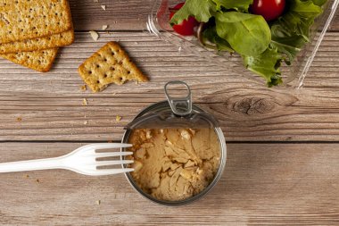 Tabakta ya da ahşap masa üzerinde ton balığı parçacıkları bulunan açık ton balığı konservesi, salata, kraker ve tek kullanımlık plastik çatallı plastik konteynır içeren düz bir resim.. 