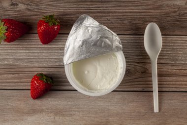 Alüminyum folyo kapağı soyulmuş bir plastik bardak düşük yağlı Yunan yoğurdu alan bir dükkanın düz görüntüsü. Plastik kaşık ve taze meyvelerle ahşap arka planda. Bir diyet fikri.