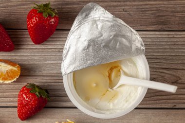 Üzerinde alüminyum folyo mührü ve mandalina dilimleri ve ahşap üzerine çilek serpiştirilmiş tek kullanımlık plastik kaşık bulunan beyaz plastik bir Yunan yoğurdu. Sağlıklı hızlı atıştırma.
