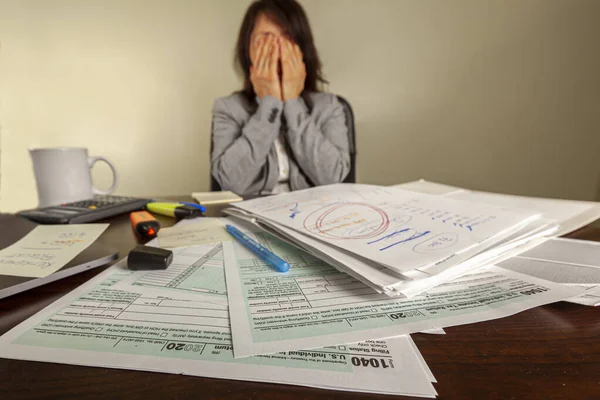 A woman is trying to file her tax return. She has piles of documents, calculator, laptop, notes and a coffee mug on her desk. She is depressed and tired of the work. She covers face with her hands