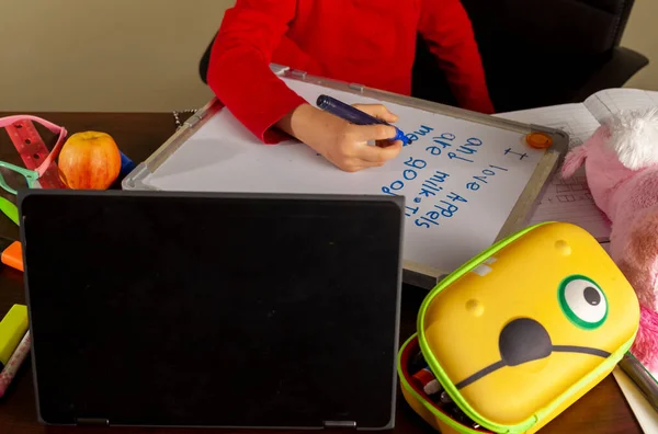 A cute little girl is writing sentences on a white board using blue marker. She is attending virtual classroom with her teacher through her laptop due to ongoing COVID 19 pandemic. This is her desk.