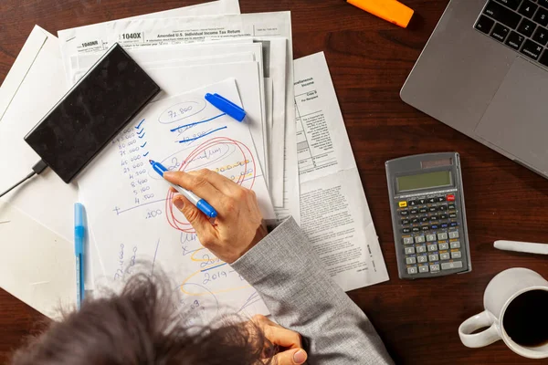 A business woman wearing formal dress is working at an office setting. She is calculating income and expenses using statements, pay slips, receipts. She uses a marker to tick items. Tax return concept