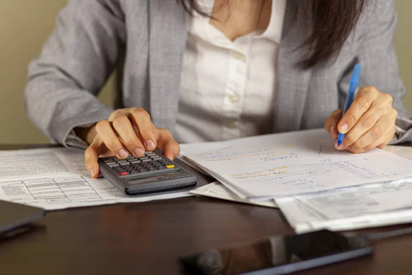 A business woman wearing formal dress is working at an office setting. She is calculating income and expenses using statements, pay slips, receipts. She uses a marker to tick items. Tax return concept