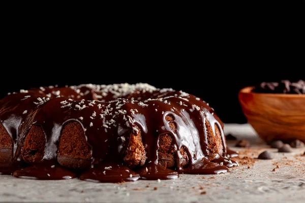 Side view of a round Chocolate cake with melted  pudding icing and chocolate chips in a bowl in the background. The decoration of this delicious cake was finished with sprinkles of shredded coconuts.