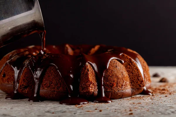 Melted hot chocolate pudding is being poured down from a metal coffee pot pitcher onto a chocolate cake as icing. It flows down and makes brown puddles on the marble surface.