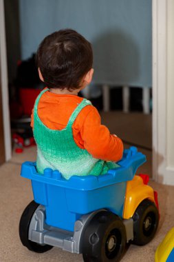 Yeşil tulumlu şirin küçük bir çocuk yatak odasındaki plastik bir oyuncak kamyonunun bagajında oturuyor. Bu oyuncakta çok eğleniyor..