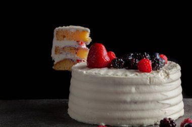 Yuvarlak bir pasta resmi, kremalı beyaz krema ve dekoratif malzemeler olarak kullanılan böğürtlen çeşitleri. Koyu gri granit arka planda servis edilmiş. Kenarında bir kesik var.