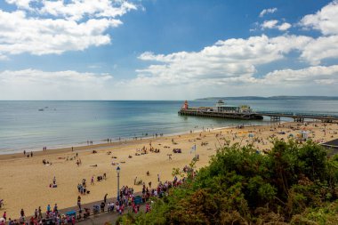 Bournemouth sahili ve rıhtımının yanı sıra sakin Atlantik okyanusu ve mavi gökyüzü manzarası. Gökyüzü, doğu uçurumlarından kuşbakışı görüntü, önünde yeşil ağaçlar var. İnsanlar kıyıda yürüyor.