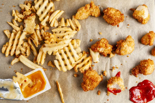 flat lay image of a messy food with waffle fries and chicken nuggets on baking paper together with ketchup and other sauces and dips. Some are eaten others dipped. A messy junk food scene.