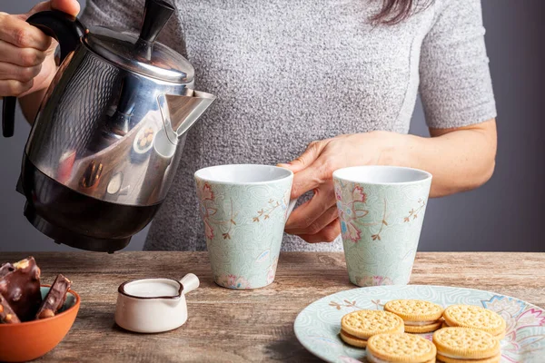 a tea time or coffee time concept with sandwich cookies, bars of chocolate aesthetic ceramic mug and plates as well as a mini creamer pitcher on wooden table. A woman is pouring hot drink into mug.