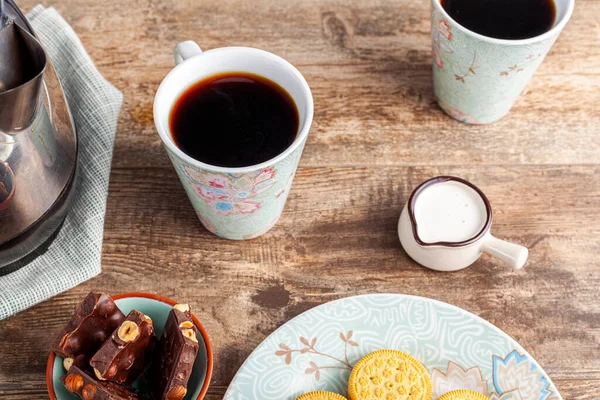 a tea time or coffee time concept with sandwich cookies, bars of chocolate aesthetic ceramic mug and plates as well as a mini creamer pitcher on wooden table.
