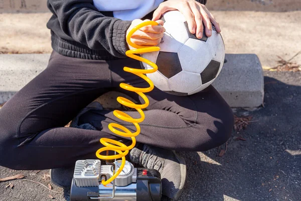 Close up image showing a caucasian woman holding a soccer ball and inserting needle bit at the end of the yellow coiled tubing attached to 12v car tire inflator to pump in air to the ball.