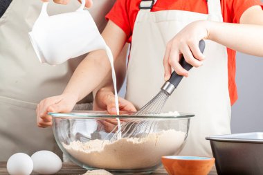 Anne ve kızı mutfak konseptinde birlikte yemek pişirirken elinde çırpı tutan kız ve sürahiden karışıma dikkatle süt döken kadın. Eğlenceli bir kooperatif aktivite.