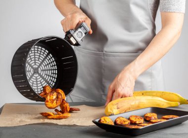 Beyaz bir kadın taze yapılmış muz cipslerini (Platanos Maduros) soğutmak için pişirme kağıdına döküyor. Kızartma sepetinin sapını tutuyor. Kızartma makinesi normal kaloriye kıyasla düşük kalori üretiyor..