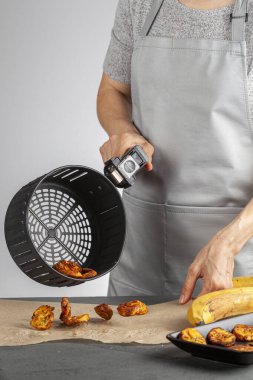 Beyaz bir kadın taze yapılmış muz cipslerini (Platanos Maduros) soğutmak için pişirme kağıdına döküyor. Kızartma sepetinin sapını tutuyor. Kızartma makinesi normal kaloriye kıyasla düşük kalori üretiyor..