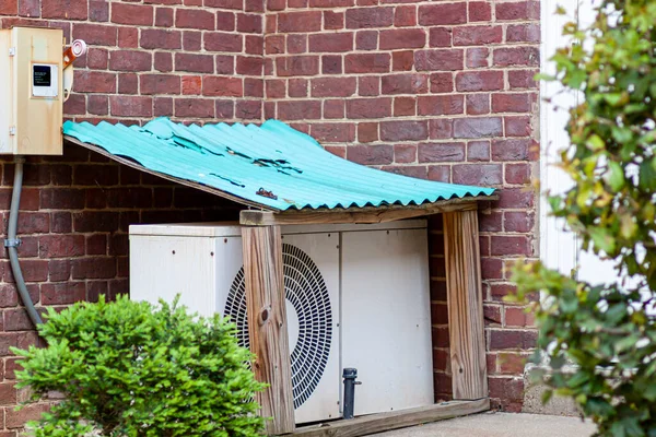 An outdoor air conditioner AC unit placed on the ground at a garden within a small protective utility shed with corrugated painted metal roof. This provides weather protection for the electrical unit.