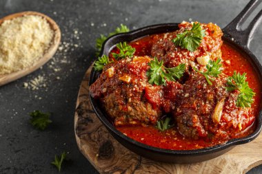 Dökme demir tavada cızırdayan domates soslu geleneksel İtalyan köftelerini yakından izleyin. Tava ahşap, maydanoz ve rendelenmiş parmesan peynirinin üzerine oturur.  