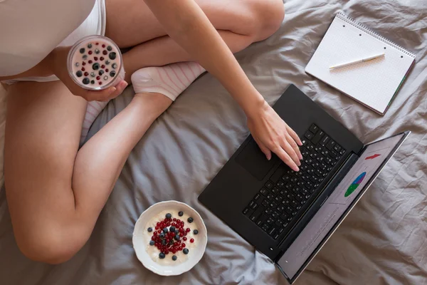 Felső shot nő láb, a laptop, a joghurt és a notebook a szürke takaró — Stock Fotó