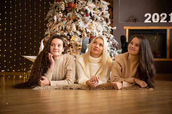 Tres hermosas y lindas chicas tumbadas cerca del árbol de Navidad, vestidas con cálidos pullovers, sonriendo y esperando las vacaciones de Navidad, Feliz Año Nuevo 2021 — Foto de Stock