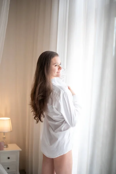 Morena en camisa de hombre. Vista trasera de la joven mujer mirando por la ventana —  Fotos de Stock