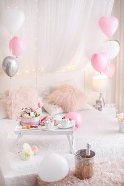 Çiçekler ve tatlılar beyaz masada ve balonlar yatağın üstünde. Sevgililer günü ya da 8 Mart ya da anneler günü ya da doğum günü hediyesi.