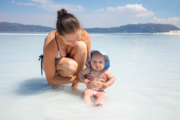 Young woman in swim suit and her daughter little baby girl are having fun on white clay of turquoise crater lake Salda Golu, Turkey