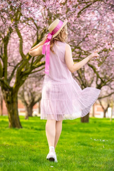 Gyönyörű nő szalmakalapban sétál egy tavaszi kertben virágzó cseresznyefákkal — Stock Fotó