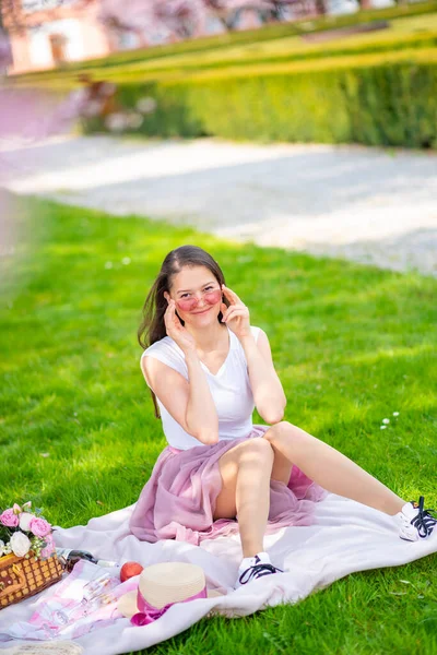 Piękna kobieta w słomkowym kapeluszu pod kwitnącymi wiśniami w miejscu picnik. Dziewczyna w różowej długiej spódnicy w wiosennym ogrodzie — Zdjęcie stockowe
