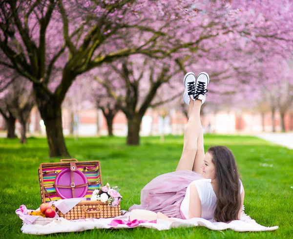 Mulher bonita no chapéu de palha abaixo de árvores de cereja florescentes no lugar de picnik. Menina vestindo uma saia longa rosa no jardim da primavera — Fotografia de Stock