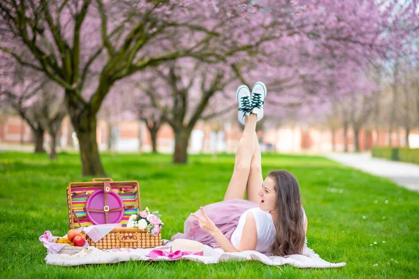 Mulher bonita no chapéu de palha abaixo de árvores de cereja florescentes no lugar de picnik. Menina vestindo uma saia longa rosa no jardim da primavera — Fotografia de Stock