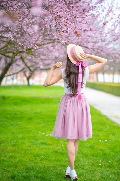 Wanita cantik dengan topi jerami berjalan di taman musim panas dengan pohon ceri mekar. Gadis mengenakan rok merah muda — Stok Foto