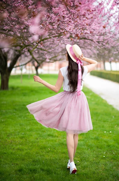 Wanita cantik dengan topi jerami berjalan di taman musim panas dengan pohon ceri mekar. Gadis mengenakan rok merah muda — Stok Foto