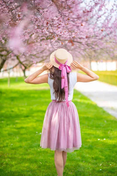Gyönyörű nő szalmakalapban sétál egy nyári kertben virágzó cseresznyefákkal. Rózsaszín szoknyás lány — Stock Fotó