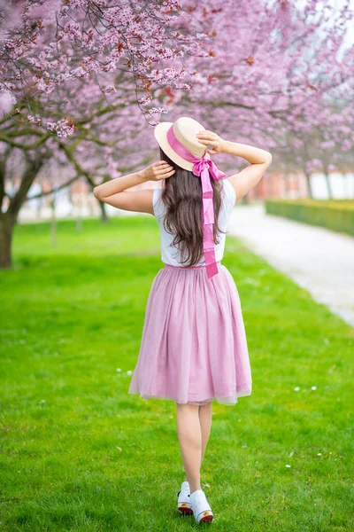 Wanita cantik dengan topi jerami berjalan di taman musim panas dengan pohon ceri mekar. Gadis mengenakan rok merah muda — Stok Foto