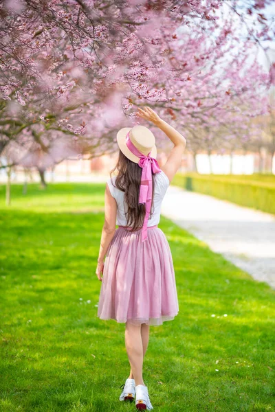 Gyönyörű nő szalmakalapban sétál egy nyári kertben virágzó cseresznyefákkal. Rózsaszín szoknyás lány — Stock Fotó