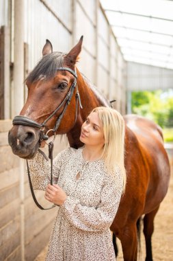 Uzun saçlı, sarışın, açık renk ahırda kahverengi atıyla poz veren genç bir atlı kadın, Sibirya, Rusya