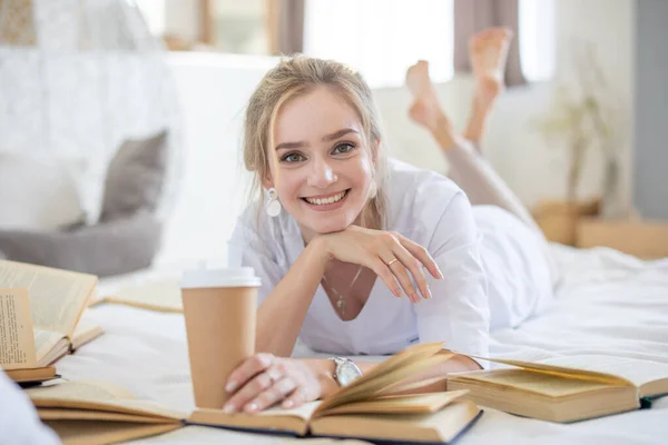 Mooie sensuele jonge vrouw met blond haar dragen van een wit shirt boeken lezen en het drinken van koffie op het bed in modern huis — Stockfoto