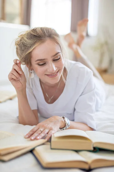 Красивая чувственная молодая женщина с светлыми волосами в белой рубашке читает книги на кровати в современном доме — стоковое фото