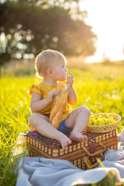 Mała dziewczynka je świeże bagietki i owoce na pikniku o zachodzie słońca w przyrodzie. — Zdjęcie stockowe