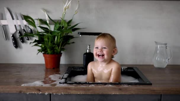 Evdeki mutfak lavabosunda su ve köpükle oynayan tatlı, mutlu bir kız. — Stok video