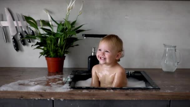 Evdeki mutfak lavabosunda su ve köpükle oynayan tatlı, mutlu bir kız. — Stok video