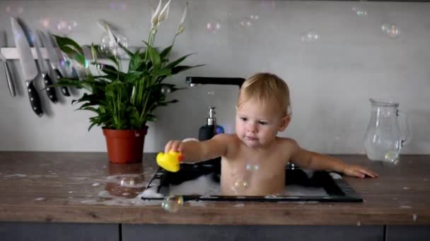 Mignon bébé fille heureuse avec jouer avec de l'eau et de la mousse dans un évier de cuisine à la maison — Video