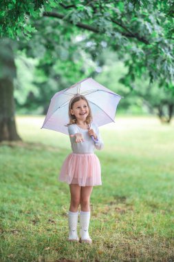 Küçük kız şemsiyesinin altındaki yağmuru yakalamak için iki eliyle gülüyor ve uzanıyor. Kaygısız ve mutlu bir hareket anı ve açık hava oyunu. Yüksek kalite fotoğraf