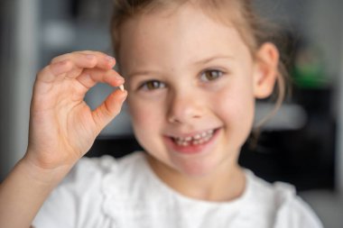 Süt dişini parmaklarının arasında tutan gülümseyen bir kızın portresi. Elinde tuttuğu dişe odaklan, önemli bir çocukluk dönüm noktasını ve neşeli deneyimi sembolize ediyor..
